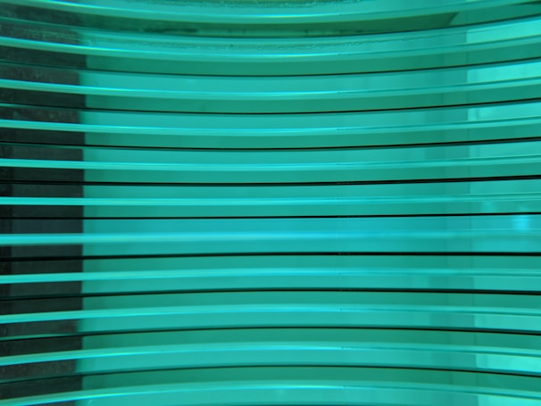 Several horizontal turquoise glass panes are stacked in parallel, creating a layered and reflective surface. The light passing through the glass gives it a luminous and modern appearance.