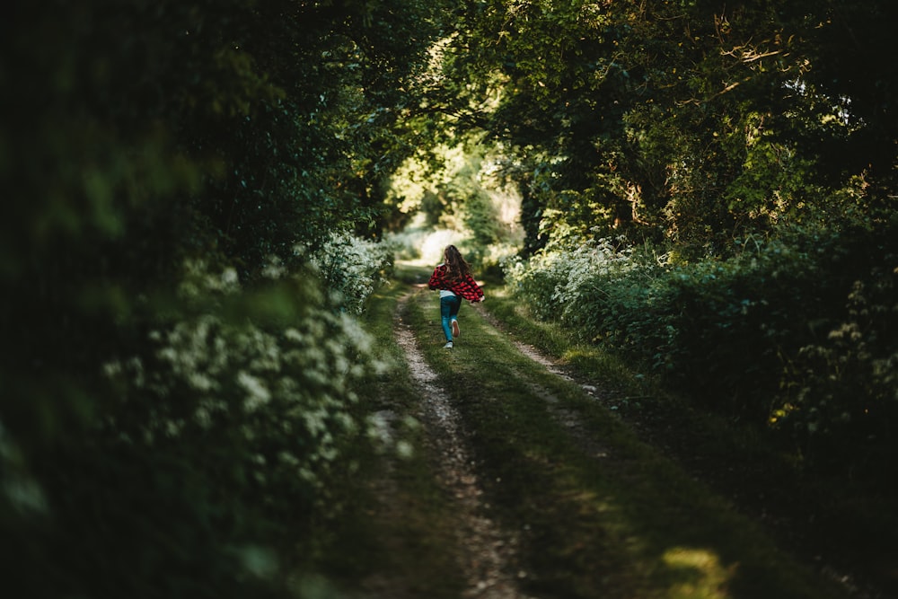 woman in red shirt and green pants walking on grass road between trees during daytime