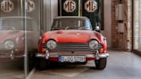 A vintage red sports car is parked indoors, reflecting off a glass surface to its left. The car features classic design elements like round headlights and chrome details. Behind it, the MG logo is prominently displayed on a green backdrop. The surroundings include visible brickwork and industrial-style elements.