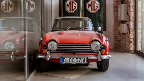 A vintage red sports car is parked indoors, reflecting off a glass surface to its left. The car features classic design elements like round headlights and chrome details. Behind it, the MG logo is prominently displayed on a green backdrop. The surroundings include visible brickwork and industrial-style elements.