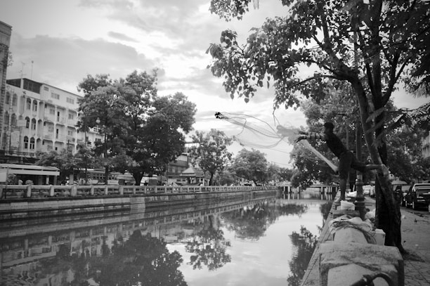 A black and white photograph captures a person casting a fishing net over a river or canal. Trees and buildings line the waterway, and the reflection of the surrounding environment is visible on the water's surface. The scene appears to be in an urban area, with a walkway and parked vehicles visible on one side.