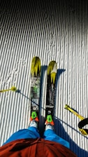 Two skis with bright yellow and green patterns are positioned on a neatly groomed, snow-covered surface. A skier's legs dressed in blue pants and red jacket are visible, along with ski poles on either side.