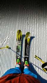 Two skis with bright yellow and green patterns are positioned on a neatly groomed, snow-covered surface. A skier's legs dressed in blue pants and red jacket are visible, along with ski poles on either side.