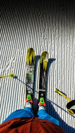 Two skis with bright yellow and green patterns are positioned on a neatly groomed, snow-covered surface. A skier's legs dressed in blue pants and red jacket are visible, along with ski poles on either side.