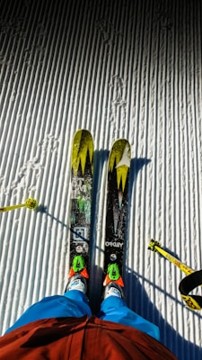 Two skis with bright yellow and green patterns are positioned on a neatly groomed, snow-covered surface. A skier's legs dressed in blue pants and red jacket are visible, along with ski poles on either side.