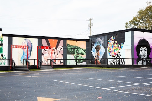 A series of colorful murals adorns the side of a building. The artwork ranges from abstract patterns to portraits, including depictions of people and a variety of vibrant designs. The building has a flat black roof and is adjacent to an empty parking lot. The setting appears to be outdoors in an urban environment with some grass and trees visible.