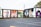A series of colorful murals adorns the side of a building. The artwork ranges from abstract patterns to portraits, including depictions of people and a variety of vibrant designs. The building has a flat black roof and is adjacent to an empty parking lot. The setting appears to be outdoors in an urban environment with some grass and trees visible.