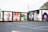 A series of colorful murals adorns the side of a building. The artwork ranges from abstract patterns to portraits, including depictions of people and a variety of vibrant designs. The building has a flat black roof and is adjacent to an empty parking lot. The setting appears to be outdoors in an urban environment with some grass and trees visible.