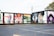 A series of colorful murals adorns the side of a building. The artwork ranges from abstract patterns to portraits, including depictions of people and a variety of vibrant designs. The building has a flat black roof and is adjacent to an empty parking lot. The setting appears to be outdoors in an urban environment with some grass and trees visible.