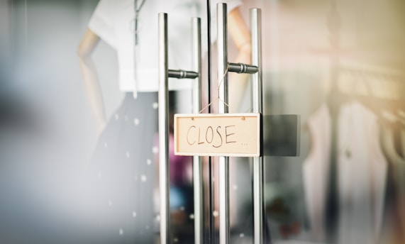 A sign hanging on glass doors displays the word 'CLOSE' in bold, with mannequins and clothing visible in the blurred background.