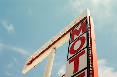 A vintage motel sign with large red letters spelling out 'MOTEL' is prominently placed against a clear blue sky. The sign features a decorative border with light bulbs and a tall, narrow design.