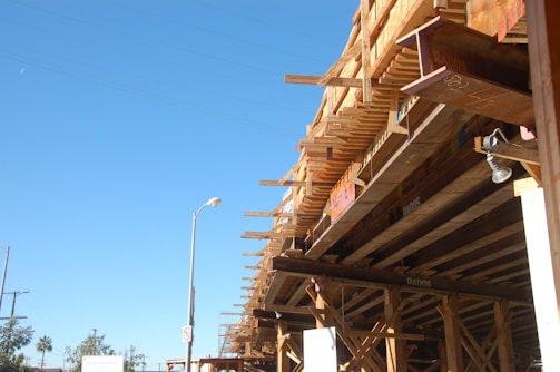 A construction site features a large wooden structure under development, supported by beams and scaffolding. The sky is clear and blue, with a street lamp and a few trees visible in the distance.