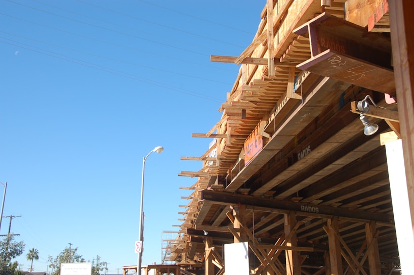 A construction site features a large wooden structure under development, supported by beams and scaffolding. The sky is clear and blue, with a street lamp and a few trees visible in the distance.