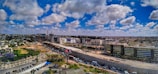 A wide urban landscape featuring expansive infrastructure development, with a highway dividing the image. Modern buildings line one side, while construction is visible in the distance. Fluffy clouds are scattered across a deep blue sky, and vehicles are moving on the road.