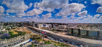 A wide urban landscape featuring expansive infrastructure development, with a highway dividing the image. Modern buildings line one side, while construction is visible in the distance. Fluffy clouds are scattered across a deep blue sky, and vehicles are moving on the road.