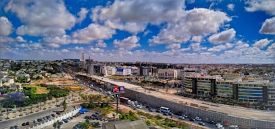 A wide urban landscape featuring expansive infrastructure development, with a highway dividing the image. Modern buildings line one side, while construction is visible in the distance. Fluffy clouds are scattered across a deep blue sky, and vehicles are moving on the road.