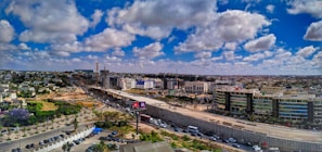 A wide urban landscape featuring expansive infrastructure development, with a highway dividing the image. Modern buildings line one side, while construction is visible in the distance. Fluffy clouds are scattered across a deep blue sky, and vehicles are moving on the road.