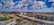A wide urban landscape featuring expansive infrastructure development, with a highway dividing the image. Modern buildings line one side, while construction is visible in the distance. Fluffy clouds are scattered across a deep blue sky, and vehicles are moving on the road.