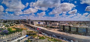 A wide urban landscape featuring expansive infrastructure development, with a highway dividing the image. Modern buildings line one side, while construction is visible in the distance. Fluffy clouds are scattered across a deep blue sky, and vehicles are moving on the road.