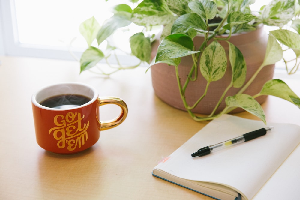 Schwarzer einziehbarer Stift auf aufgeschlagenem Buch neben rot-weißer Go Get'em-bedruckter Kaffeetasse