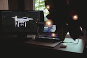 A workspace features a laptop and a monitor. The laptop screen displays a video editing software interface with a clip of a person swimming. The monitor on the left shows an image of a drone flying over a forest. A plant with decorative lights partially obscures the scene from the right.