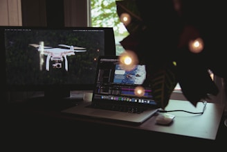 A workspace features a laptop and a monitor. The laptop screen displays a video editing software interface with a clip of a person swimming. The monitor on the left shows an image of a drone flying over a forest. A plant with decorative lights partially obscures the scene from the right.