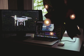 A workspace features a laptop and a monitor. The laptop screen displays a video editing software interface with a clip of a person swimming. The monitor on the left shows an image of a drone flying over a forest. A plant with decorative lights partially obscures the scene from the right.