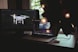 A workspace features a laptop and a monitor. The laptop screen displays a video editing software interface with a clip of a person swimming. The monitor on the left shows an image of a drone flying over a forest. A plant with decorative lights partially obscures the scene from the right.
