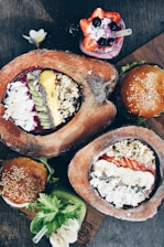 A variety of colorful food items presented on a wooden surface, including smoothie bowls garnished with fresh fruits like kiwi, strawberries, and bananas, topped with nuts and seeds. Accompanying the bowls are two burgers with sesame seed buns and a vibrant green smoothie with a banana slice.