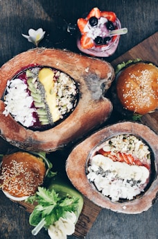 A variety of colorful ood items presented on a wooden surface, including smoothie bowls garnished with fresh fruits like kiwi, strawberries, and bananas, topped with nuts and seeds. Accompanying the bowls are two burgers with sesame seed buns and a vibrant green smoothie with a banana slice.