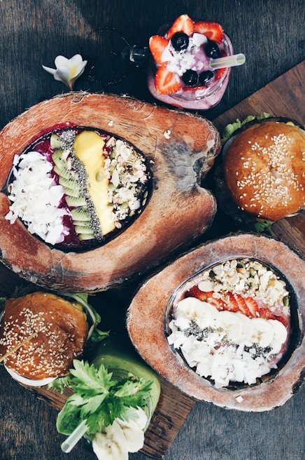 A variety of colorful food items presented on a wooden surface, including smoothie bowls garnished with fresh fruits like kiwi, strawberries, and bananas, topped with nuts and seeds. Accompanying the bowls are two burgers with sesame seed buns and a vibrant green smoothie with a banana slice.