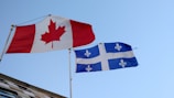 Two flags are waving against a clear blue sky. The first flag has a red maple leaf on a white background with red bars on each side, representing Canada. The second flag is blue with a white cross and four white fleurs-de-lis, representing Quebec.