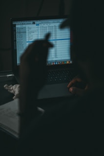A dimly lit setting featuring a person working on a laptop, with a screen displaying a list of media files. The person's silhouette is visible, suggesting focused attention on the screen. There are some notes taken on a notepad alongside the computer.