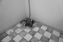 A black and white image featuring a coiled hose reel with a cable, placed near a corner of a room with tiled flooring. The tiles have a geometric pattern and the walls are plain.