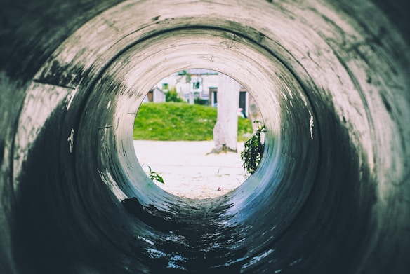 A large concrete pipe with a view looking out towards an outdoor scene. The interior of the pipe is smooth and cylindrical, leading to an opening where greenery and part of a tree trunk are visible. Sunlight illuminates part of the pipe's inside and the surrounding outside area.