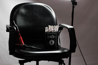 A black salon chair with armrests is set against a neutral background. Positioned on the seat, a small black bag is visible, containing various hair styling tools and clips. To the left, a red and black curling iron lies across the chair, and a hairbrush rests nearby. A soft light source illuminates the chair, creating subtle shadows.