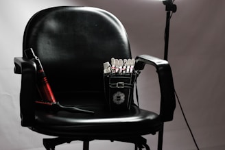 A black salon chair with armrests is set against a neutral background. Positioned on the seat, a small black bag is visible, containing various hair styling tools and clips. To the left, a red and black curling iron lies across the chair, and a hairbrush rests nearby. A soft light source illuminates the chair, creating subtle shadows.