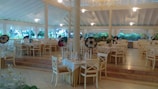 A spacious dining area features elegant wooden chairs and tables set with white tablecloths. Sombreros are placed on some chairs, adding a festive touch. Large windows allow natural light to illuminate the space, with lush green foliage visible outside. Ceiling fans hang from the high, vaulted ceiling, contributing to a tropical and airy atmosphere.