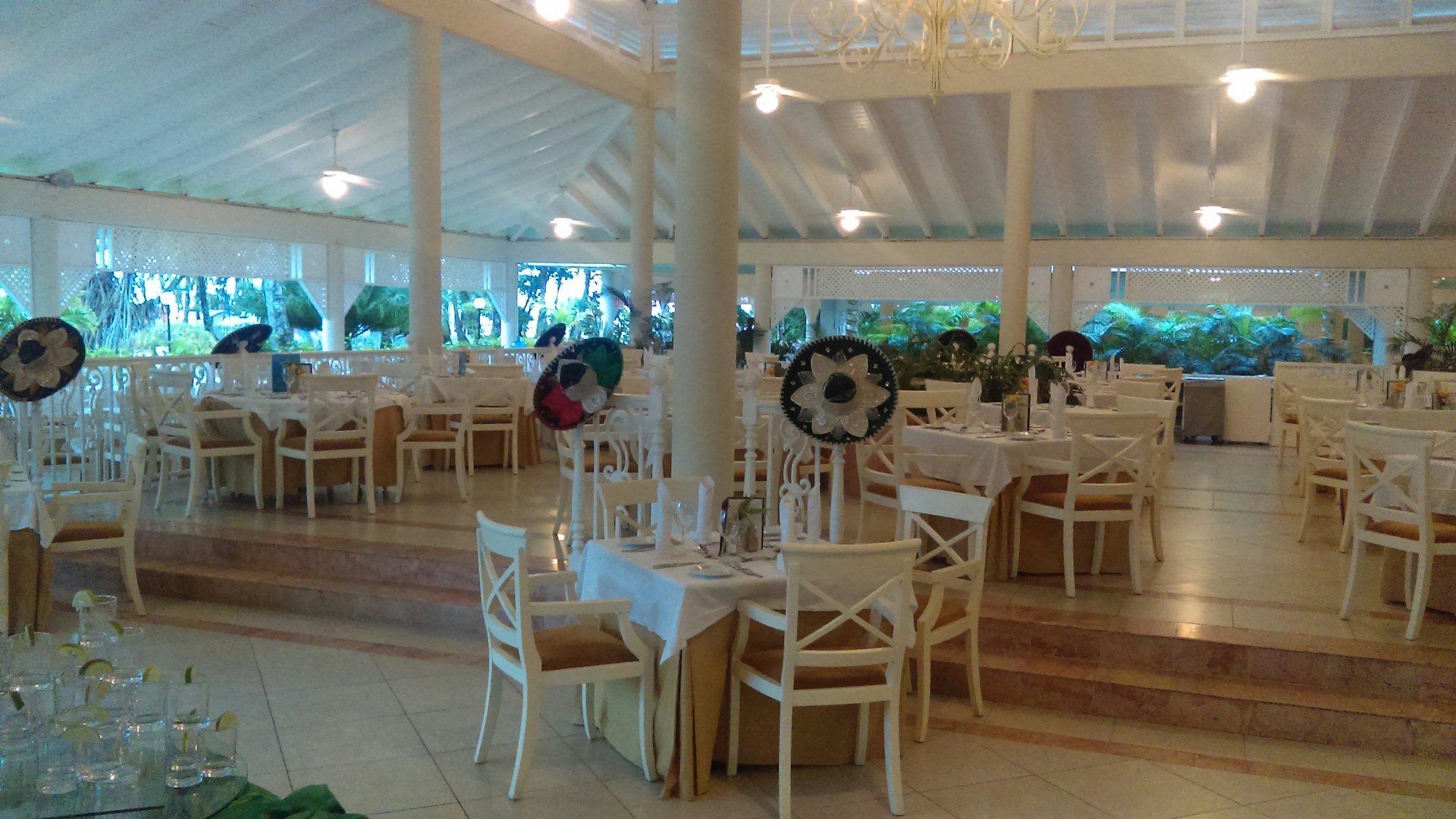 A spacious dining area features elegant wooden chairs and tables set with white tablecloths. Sombreros are placed on some chairs, adding a festive touch. Large windows allow natural light to illuminate the space, with lush green foliage visible outside. Ceiling fans hang from the high, vaulted ceiling, contributing to a tropical and airy atmosphere.