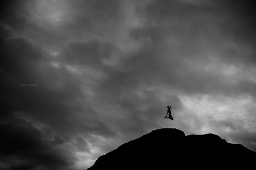 Silhouette of a person meditating in a seated position against a dramatic cloudy sky backdrop. The person appears to be levitating above a rocky terrain, adding a sense of mystery and contemplation.