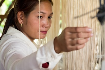 A woman wearing a white shirt is engaged in a weaving activity, carefully handling a thread from a loom. Her focused expression indicates concentration on the task at hand, surrounded by strands of light-colored thread.