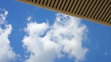 White clouds drift across a vibrant blue sky. An overhanging structure with a slatted design cuts across the top of the image, adding a geometric contrast to the softness of the clouds.
