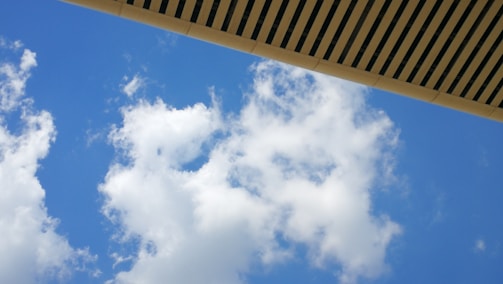 White clouds drift across a vibrant blue sky. An overhanging structure with a slatted design cuts across the top of the image, adding a geometric contrast to the softness of the clouds.