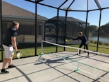 Two people are playing pickleball on a concrete court enclosed by a screened structure. They are dressed in casual athletic attire. One is about to hit a ball with a paddle, while the other watches attentively. The court is outdoors, with grass and a body of water visible in the background under a clear blue sky.