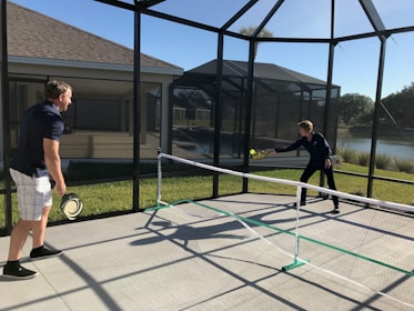 Two people are playing pickleball on a concrete court enclosed by a screened structure. They are dressed in casual athletic attire. One is about to hit a ball with a paddle, while the other watches attentively. The court is outdoors, with grass and a body of water visible in the background under a clear blue sky.