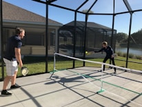 Two people are playing pickleball on a concrete court enclosed by a screened structure. They are dressed in casual athletic attire. One is about to hit a ball with a paddle, while the other watches attentively. The court is outdoors, with grass and a body of water visible in the background under a clear blue sky.