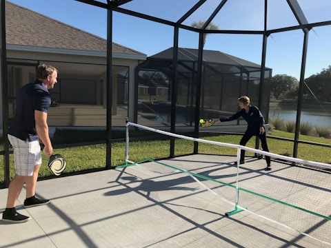 Two people are playing pickleball on a concrete court enclosed by a screened structure. They are dressed in casual athletic attire. One is about to hit a ball with a paddle, while the other watches attentively. The court is outdoors, with grass and a body of water visible in the background under a clear blue sky.