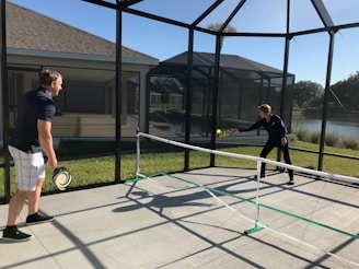 Two people are playing pickleball on a concrete court enclosed by a screened structure. They are dressed in casual athletic attire. One is about to hit a ball with a paddle, while the other watches attentively. The court is outdoors, with grass and a body of water visible in the background under a clear blue sky.