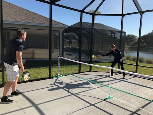 Two people are playing pickleball on a concrete court enclosed by a screened structure. They are dressed in casual athletic attire. One is about to hit a ball with a paddle, while the other watches attentively. The court is outdoors, with grass and a body of water visible in the background under a clear blue sky.
