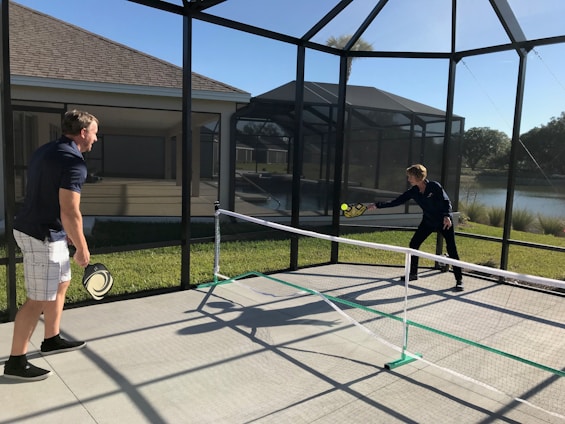Two people are playing pickleball on a concrete court enclosed by a screened structure. They are dressed in casual athletic attire. One is about to hit a ball with a paddle, while the other watches attentively. The court is outdoors, with grass and a body of water visible in the background under a clear blue sky.